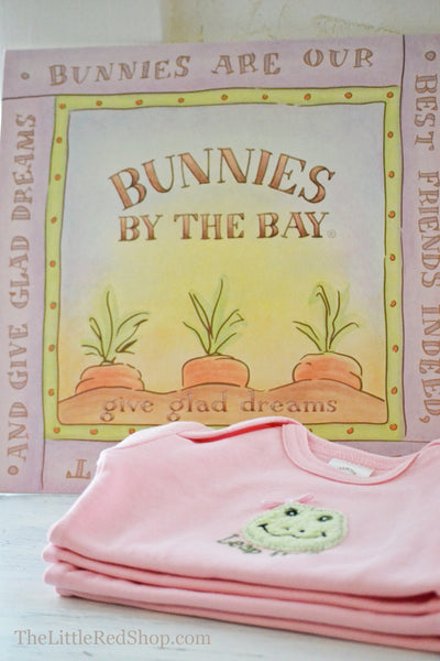 A folded stack of Bunnies by the Bay's Pink Lily Mae Leap Frog Bunsie Baby Onesie in front of Carrot Logo