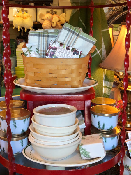 A red metal display shelf in The Little Red Shop, displaying a selection of fine curated gifts, including a woven basket filled with plaid kitchen linens, Shea Butter Soap, petite Frasier Fir hand creams, stacked cream-colored vintage ironstone bowls, platters, and stacks of Thymes Frasier Fir Gold Tin Candles with Pine Needle Design.