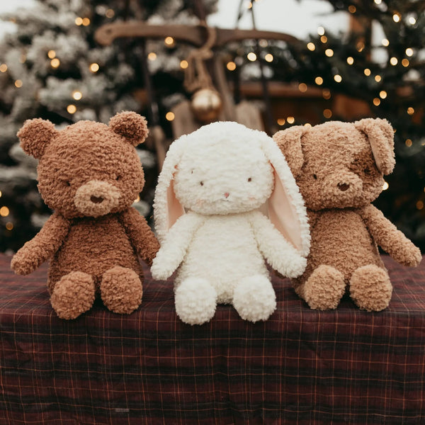 Three plush stuffed toys sit on a plaid fabric surface: a brown teddy bear, white bunny, and light brown dog with a softly blurred holiday tree and warm lights in the background.