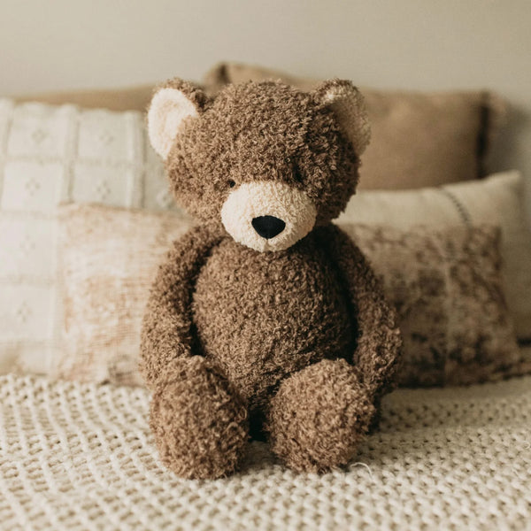 A plush teddy bear toy in a textured brown bouclé fabric, sitting on a cream knit blanket with neutral-toned pillows in the background.