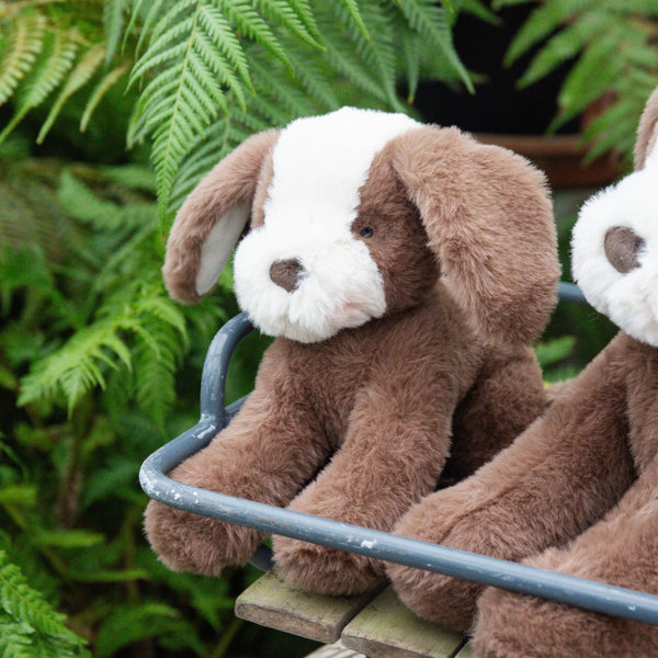 Two brown plush dogs sitting on a metal bench with green foliage in the background