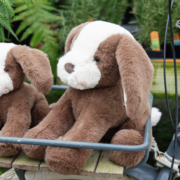 Two brown plush dogs sitting on a bench with green foliage in the background
