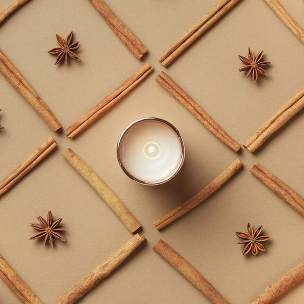 A lit white votive candle in a copper glass jar, arranged on a beige background with cinnamon sticks and star anise forming a geometric pattern.