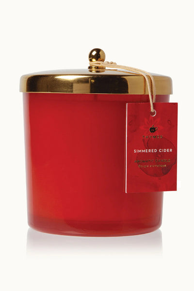 An orange-red glass jar candle with a gold metal lid and a hanging red tag that reads 'SIMMERED CIDER.'