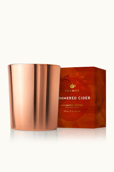 A copper-colored glass votive candle jar next to its orange and red gift box, featuring the brand name 'Thymes' and the scent 'Simmered Cider'.