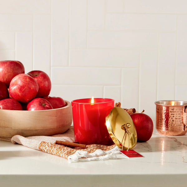 A lit red glass candle jar sits on a white countertop with a gold lid leaning beside it, surrounded by red apples, cinnamon sticks, a woven cloth, and a hammered copper mug against a white tiled wall.