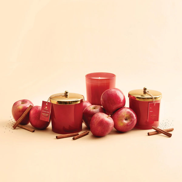 Three simmered cider scented candles in glass jars with gold lids are arranged with red apples and cinnamon sticks on a warm beige background. One candle is open, showing the wax inside.