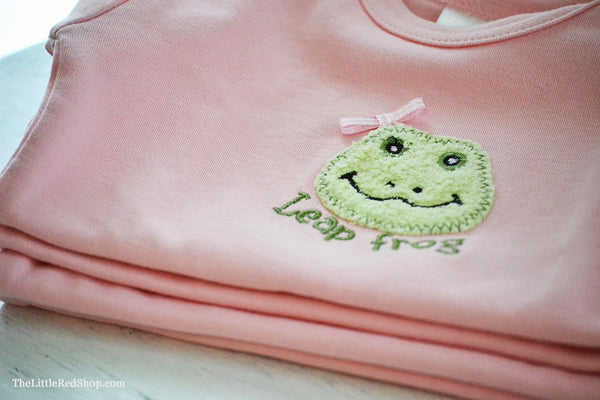 Close up of a A folded stack of Bunnies by the Bay's Pink Lily Mae Leap Frog Bunsie Baby Onesie